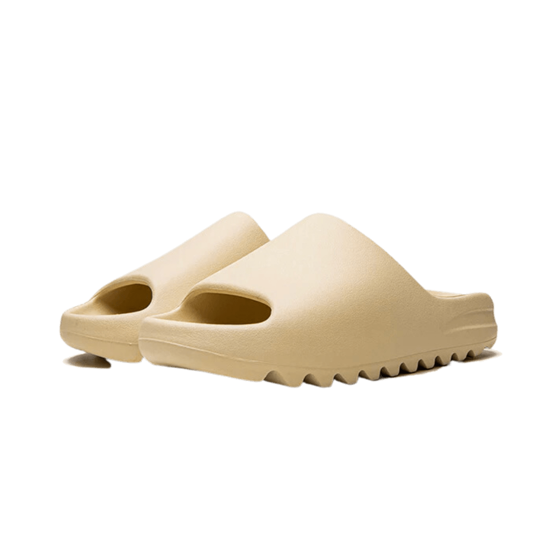 Beige Adidas Yeezy Slide Bone slippers met reliëfzool op een groene achtergrond. De schoenen zijn modern en comfortabel ontworpen voor dagelijks gebruik.