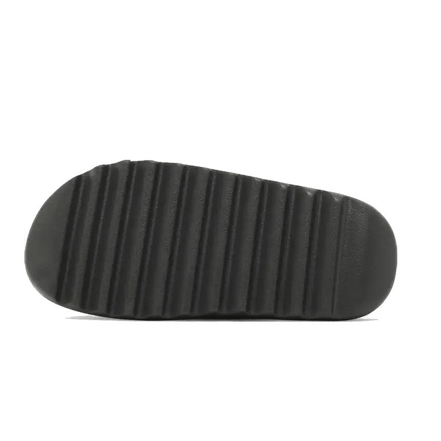 Moderne zwarte Adidas Yeezy Slide Dark Onyx slippers op een groene achtergrond.