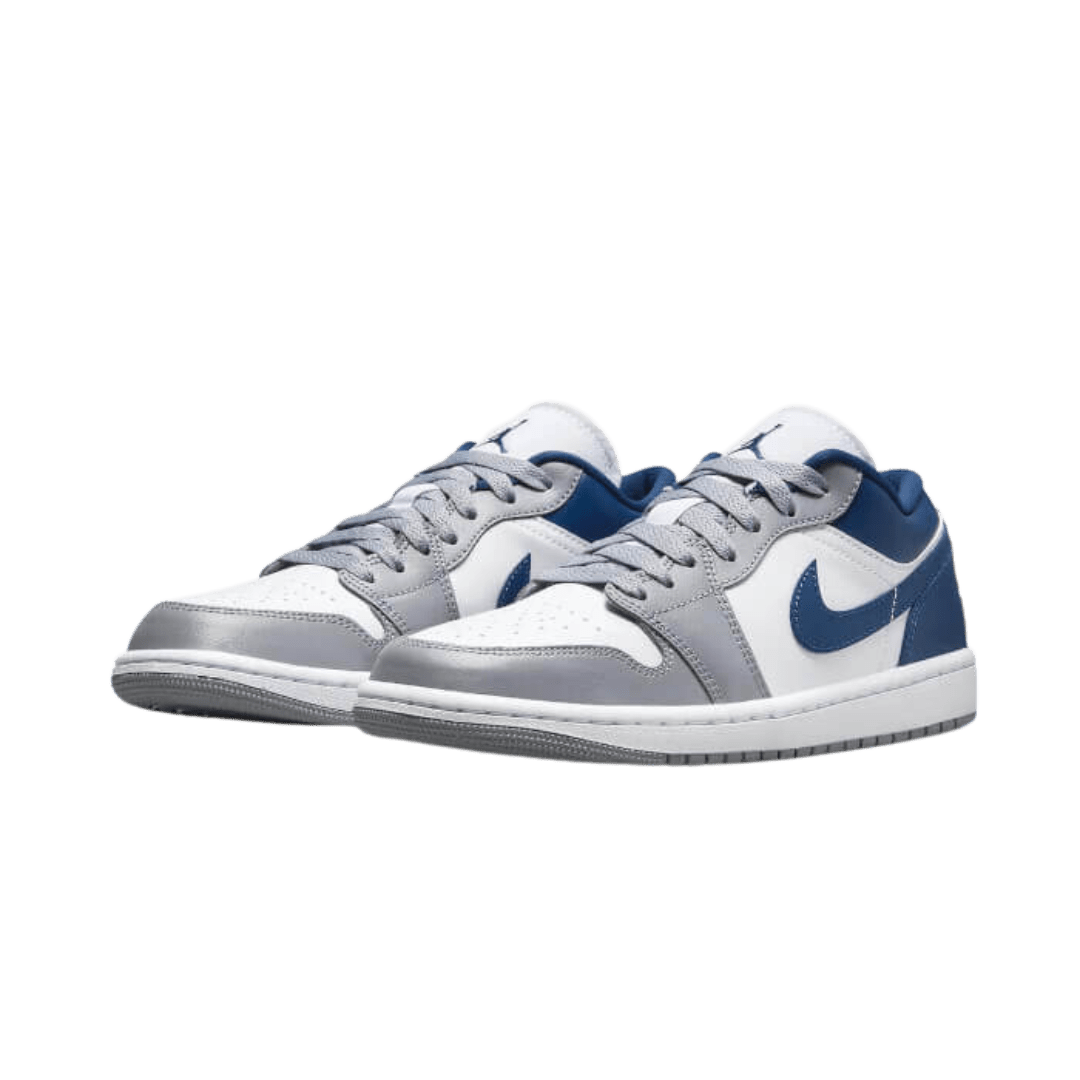Grijze en blauwe Nike Air Jordan 1 Low sneakers op een groene achtergrond. De sneakers hebben een klassiek Nike-ontwerp met een wit, grijs en blauw kleurenschema. De Nike-logo's zijn duidelijk zichtbaar op de zijkanten van de schoenen.