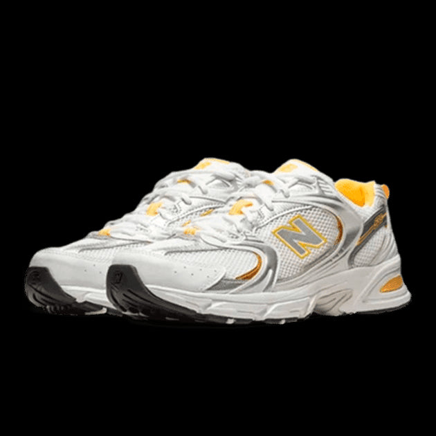 Moderne New Balance 530 sneakers in een wit, zilver en gele kleurstelling. De schoenen hebben een rubberen zool voor stabiliteit en grip, en de geperforeerde bovenlaag biedt ventilatie. Een stijlvol en comfortabel paar sportschoenen.