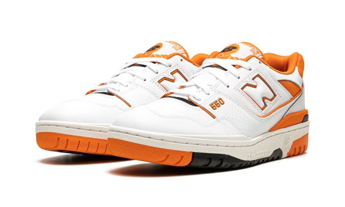 Witte en oranje New Balance 550 sneakers tegen een witte achtergrond. De sneakers hebben een klassiek ontwerp met opvallende oranje accenten en hebben een robuuste, duurzame zool.