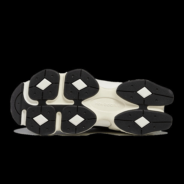 Zwarte en witte New Balance 9060 sneakers op een groene achtergrond. De zool heeft een opvallend, gedetailleerd ontwerp met geometrische vormen.