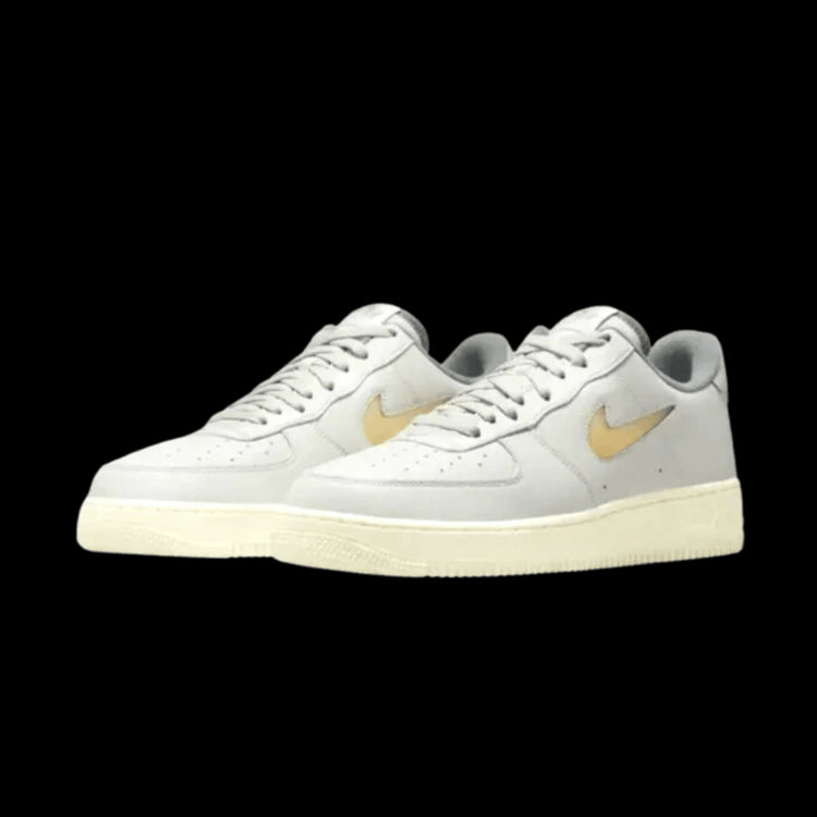 Elegant paar Nike Air Force 1 Low sneakers in lichte beige kleur met gouden accenten op een donkergroene achtergrond. De klassieke sneakers hebben een sportieve uitstraling en zijn een modetrend.