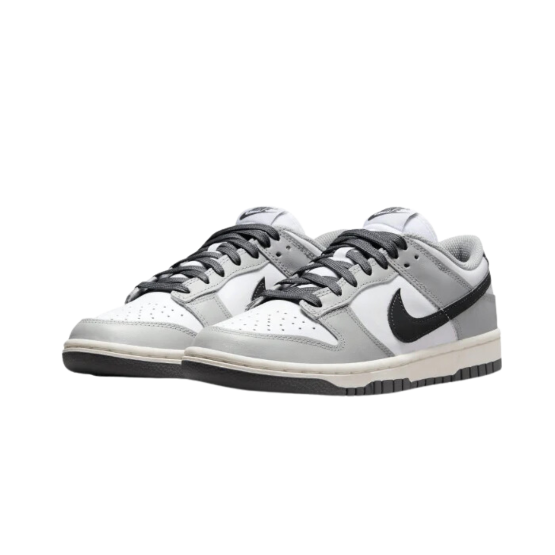 Nike Dunk Low Light Smoke Grey - Moderne sneakers met grijs en wit kleurenschema, op een zwarte achtergrond geplaatst. De schoen heeft een klassiek Dunk-design met een leren bovenkant en een rubberen zool voor duurzaamheid en grip.