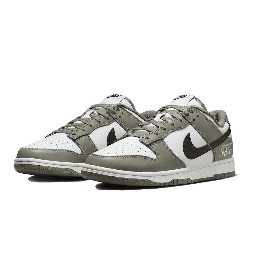 Nike Dunk Low NBA Paris sneakers op een groene achtergrond. Klassiek sportieve stijl met een tweekleurig ontwerp in grijs en wit. Kenmerkende Nike-logo's en details op de zijkanten en tong.