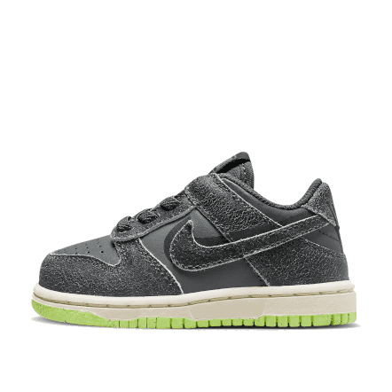Dunkelgraue Nike Dunk Low Swoosh Shadow Baby-Sneakers met lichtgroene accenten, geplaatst op een groen oppervlak.